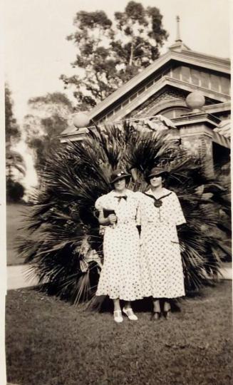 [two women dressed similarly in front of bush and building]