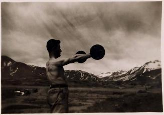 [shirtless man lifting weight in front of mountain landscape]