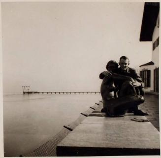 [man crouching next to a body of water with his arms around a sculpture of a woman]
