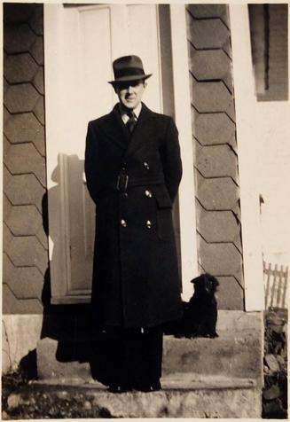 [man in dark coat and hat on steps of house with black dog]