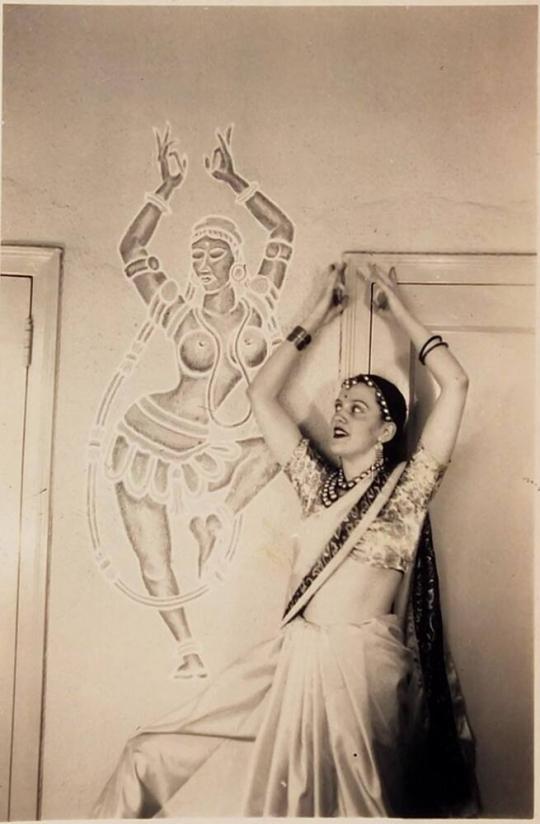 [woman in traditional Indian dress posing in front of image on wall of Indian dancer]