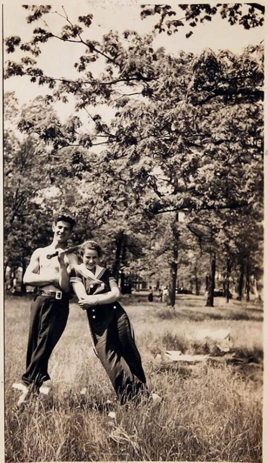 Shirtless Man And Woman Standing Leaning Against Each Other In Field With Trees In Background