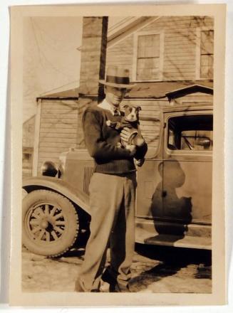 [man holding dog in front of car and house]
