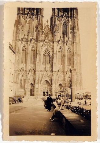 [cathedral with seated woman posing in front and two other people walking behind her]