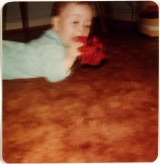 [Baby Laying on Belly on Orange Carpet Putting a Red Object in Mouth]