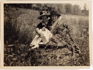 [two smiling women in a field sitting on a rock]