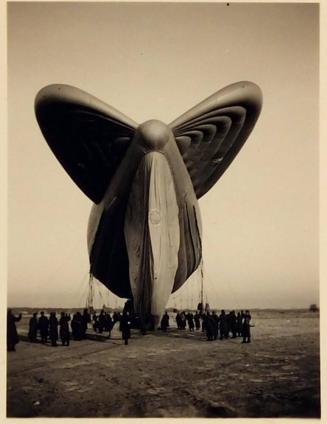 [zeppelin with large wings tied to the gournd with a crowd of people in front]