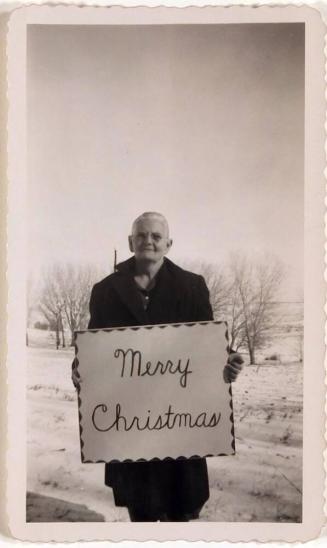[person in coat holding "Merry Christmas" sign with snowy landscape in background]