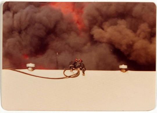 [two firefighters on top of roof with smoke and flames behind]