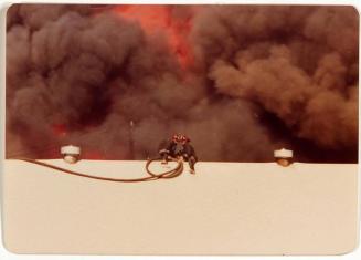 [two firefighters on top of roof with smoke and flames behind]