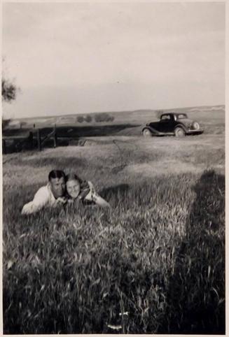 [man and woman laying in grass facing camera with car in background and shadow at right]