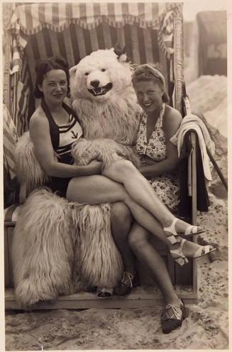 [one woman sitting on lap of person dressed as polar bear and one woman sitting next to them on beach]