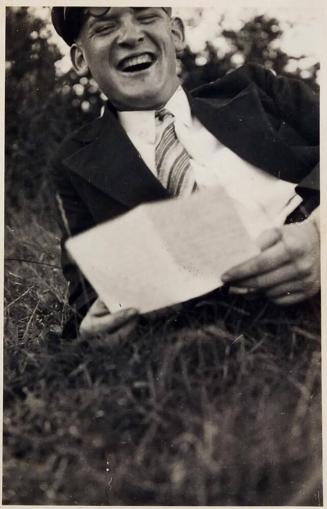 [man in suit and cap smiling with paper in hands laying in grass]