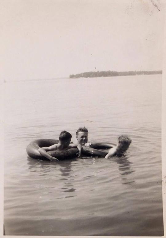 [two boys in innertubes with man holding on to innertubes in water]