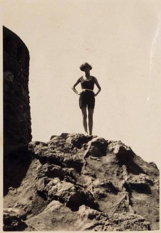 [woman standing arms akimbo on top of rock with building at left]