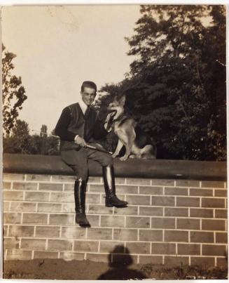 [man in riding boots holding crop sitting on wall with dog; shadow of photogropher in foreground]