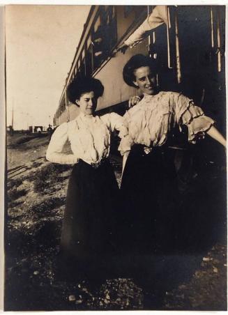 [two women in similar white blouses and long dark skirts standing in front of train]