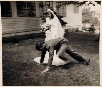 [bride standing with bouquet and groom dropping to ground on lawn]