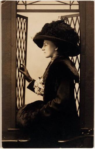 [postcard- woman in dark hat and dress sitting in window looking left]