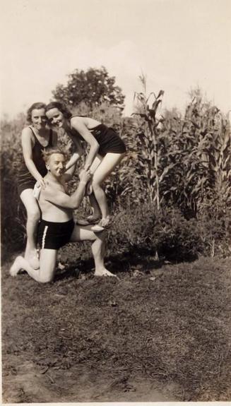 [two women standing on man in swimsuits in corn field]