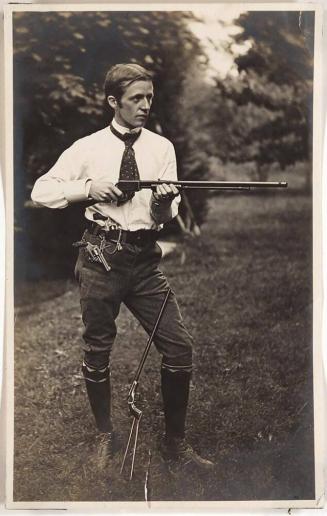 [man standing holding rifle with another rifle leaning against leg and handgun on belt]