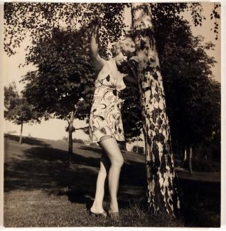 [woman in sleeveless dress leaning on trunk of tree]