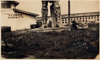[man and dog at the foot of a large statue next to a building with "FLOWER EXHIBITS" painted on the side next to a car]