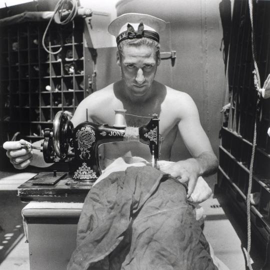 [A Royal Navy sailor on board HMS Alcantara uses a portable sewing machine to repair a signal flag during a voyage to Sierra Leone]