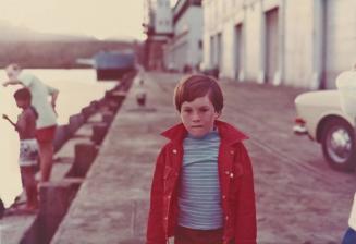 [boy with red shirt and striped shirt beneath at waterfront]