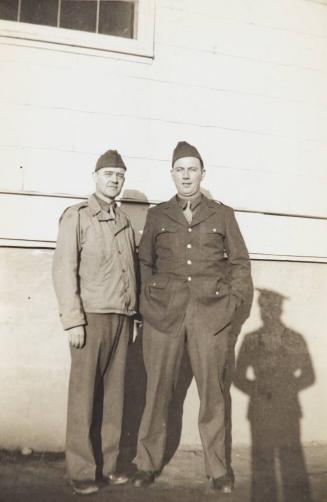 [two men in military uniform with shadow of another man at right against building]