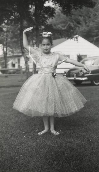 [young girl in dress with ballet shoes posing in grass with car and house in background]