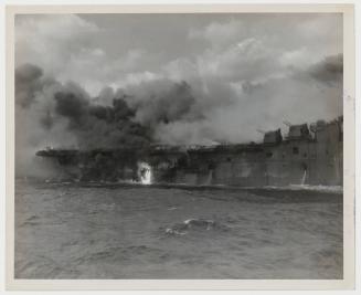 Highlighted by an area of flame, smoke spreads a choking curtain over the stern of the carrier.  Behind the pall scores of courageous men risked their lives as they exerted every effort to save their ship.