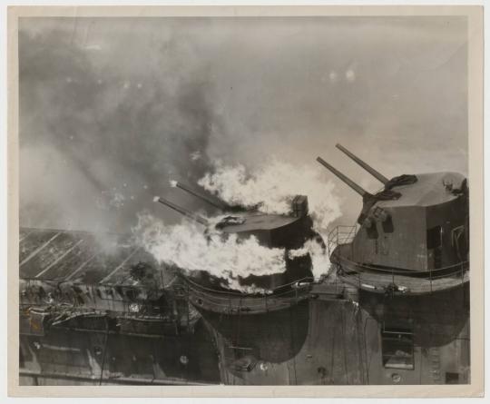 A crown of flame mantles one of the five-inch turrets on the FRANKLIN, with jets of fire flickering out of the muzzles of the twin rifles.  Crewmen aboard the cruiser SANTA FE extinguished the blaze by playing streams of water through the open hatch.