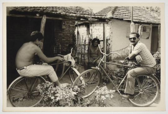 [Two Men Sitting on Bicycles' Rear Racks and a Woman Sitting on a Chair on a Porch]