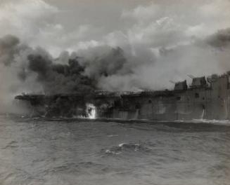 Highlighted by an area of flame, smoke spreads a choking curtain over the stern of the carrier.  Behind the pall scores of courageous men risked their lives as they exerted every effort to save their ship.