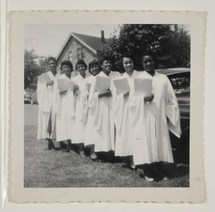 [Group of Seven Young Women in White Graduation Gowns Holding Diplomas]
