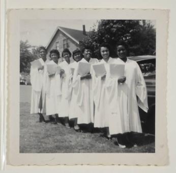 [Group of Seven Young Women in White Graduation Gowns Holding Diplomas]
