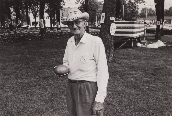 [Man in Straw Hat Holding Vegetable, Chairs, Field, Scott Orr Signs Back]